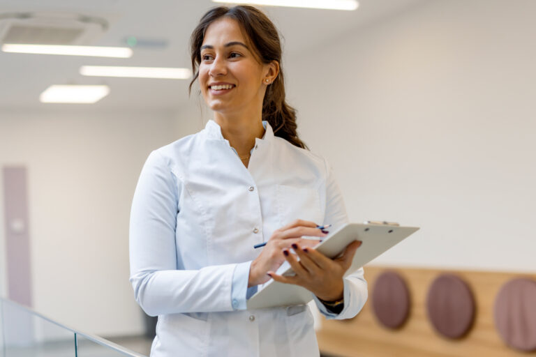 Young female doctor smiling and writing notes on a clipboard while looking away in a bright, modern hospital corridor, showcasing professionalism in patient care