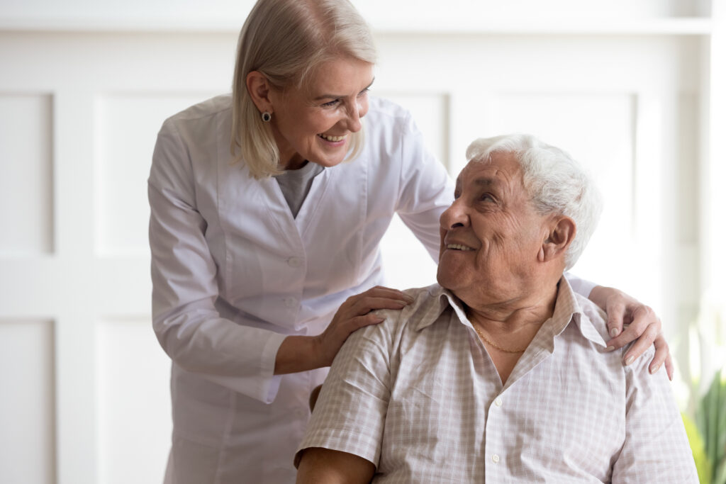 Smiling middle-aged female nurse talk take care with happy optimistic senior 80s male patient, positive woman doctor or caregiver help show support and care to mature man in retirement house