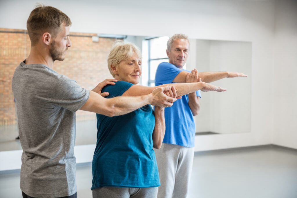 Senior people exercising at gym with trainer helping woman while doing arms workout. Old man and woman under going fitness training at rehab club.