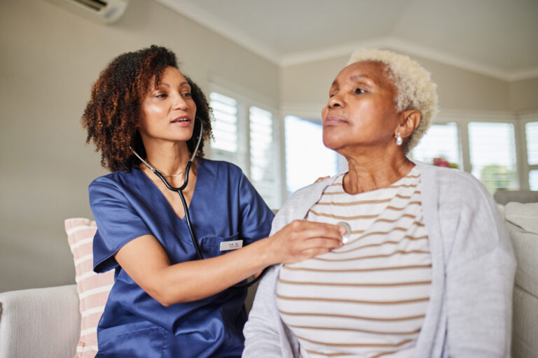 A healthcare professional listens to an elderly woman's heartbeat with a stethoscope in a bright, comfortable home setting. Focus on trust and care.