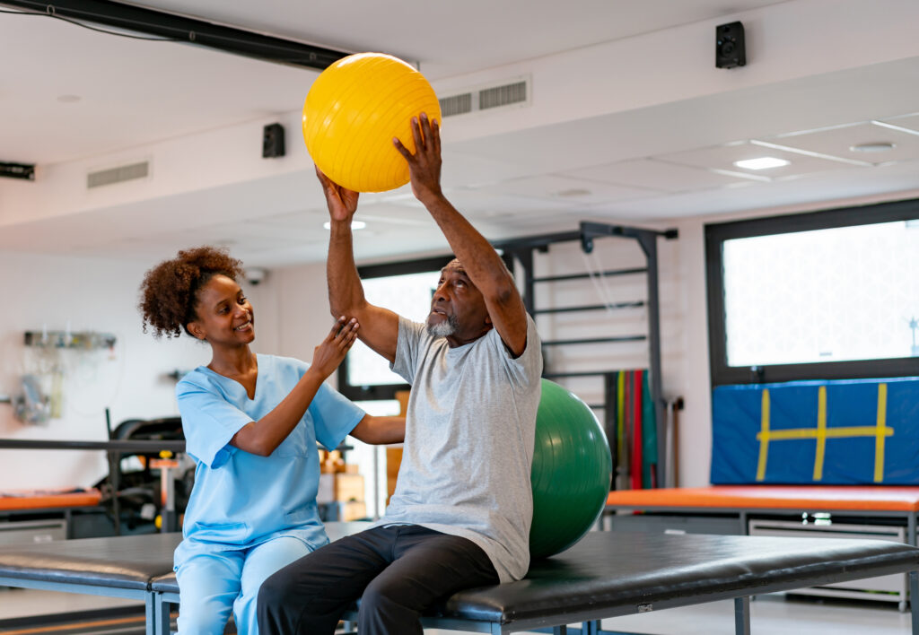 Cheerful physiotherapist helping senior black patient workout with a fitness ball at a rehabilitation center - healthcare concepts